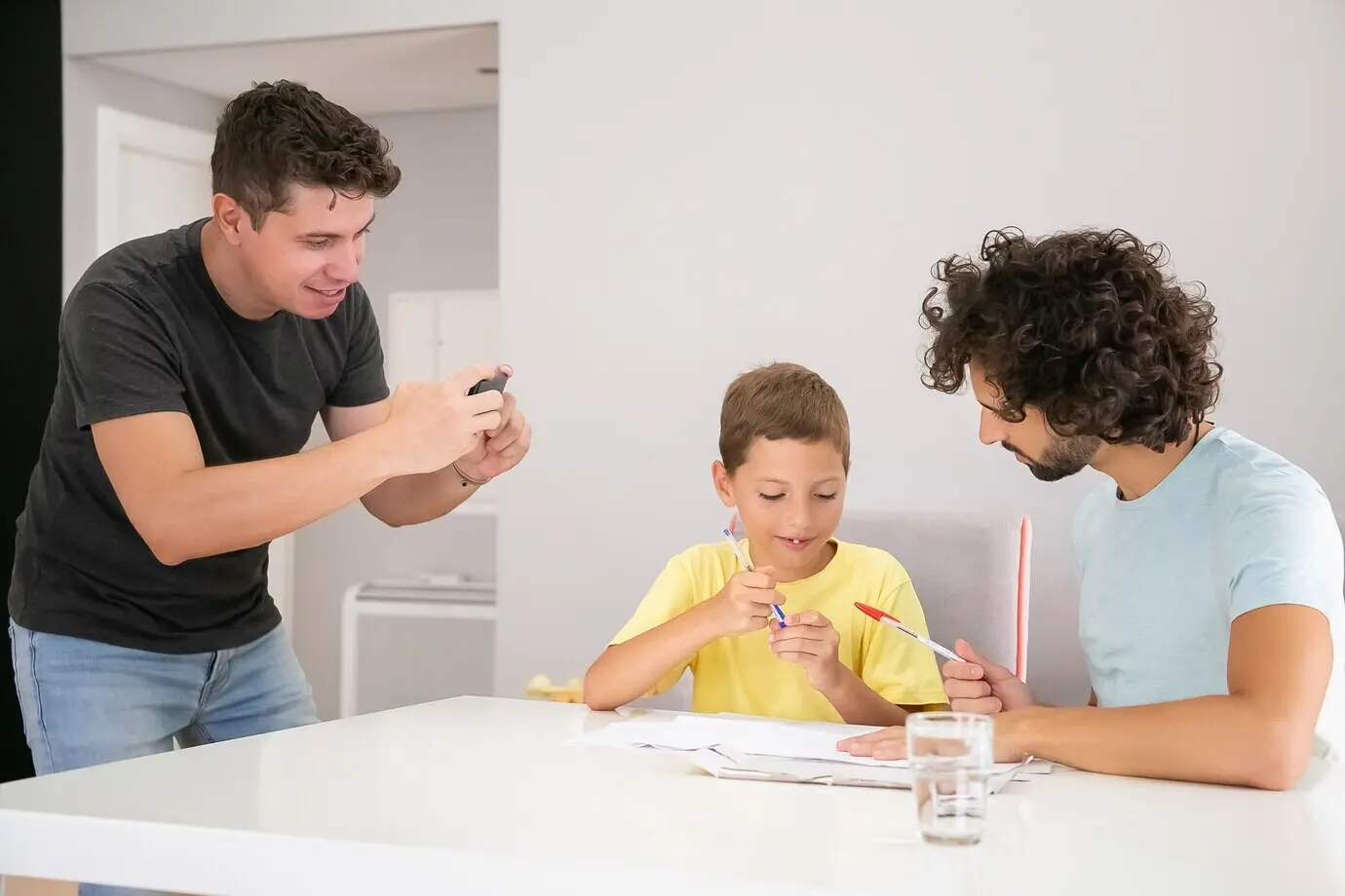 Милый, позитивный ребёнок выполняет школьное домашнее задание с помощью двух пап, пишет на листах бумаги. Мужчина фотографирует свою семью. Концепция семьи и гей-родителей.
